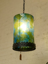 Load image into Gallery viewer, A hanging pendant lamp with a cylindrical glass shade featuring a marbled green and blue pattern, illuminated from within. The shade is finished with a black metal band at the bottom and a small tassel hangs from the chain.

