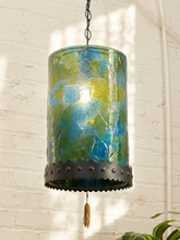 Load image into Gallery viewer, A hanging pendant lamp with a cylindrical glass shade featuring a mottled pattern of blue, green, and yellow tones, suspended by a black chain against a white brick wall.
