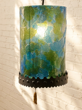 Load image into Gallery viewer, A hanging cylindrical lampshade with a marbled blue, green, and turquoise pattern, finished with a black scalloped metal band and a small tassel, shown against a white brick wall.
