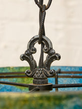 Load image into Gallery viewer, Close-up of an ornate dark metal hanging fixture with a decorative scrollwork bracket and twisted hanging chain, set against a softly blurred background with blue and green tones.
