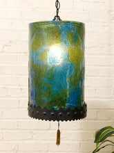 Load image into Gallery viewer, A hanging cylindrical pendant lamp with a glossy marbled green and blue glass shade, a black metal scalloped base, and a tassel, shown against a white brick wall.
