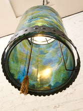 Load image into Gallery viewer, A large cylindrical pendant lamp shade made of textured, hand-blown glass with swirling green, blue, and yellow patterns, framed by a black metal band with rivets and a black scalloped rim. A gold tassel hangs from the center.
