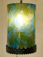 Load image into Gallery viewer, A cylindrical pendant light with a textured, hand-blown glass shade featuring a mottled pattern of green, blue, and yellow, illuminated from within. The bottom of the shade is finished with a black scalloped metal band with decorative rivets, hanging against a white brick wall.
