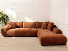 Load image into Gallery viewer, A large L-shaped sectional sofa upholstered in a warm rust-brown corduroy fabric, set against a white brick wall and light beige carpet with a green potted plant on the left.
