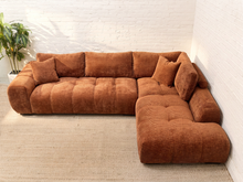 Load image into Gallery viewer, A rust-brown modular sectional sofa with tufted cushions and matching throw pillows, shown in a bright room with white brick walls and a light rug.
