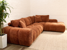 Load image into Gallery viewer, A modern L-shaped sectional sofa upholstered in a textured rust-brown corduroy fabric, styled in a minimalist living room with a white brick wall, a beige rug, and a potted green plant.
