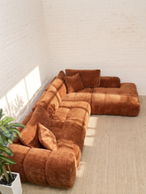 Load image into Gallery viewer, A modular L-shaped sectional sofa upholstered in a warm rust-brown bouclé fabric, styled in a minimalist room with a white brick wall, beige carpet, and a potted green plant in the foreground.
