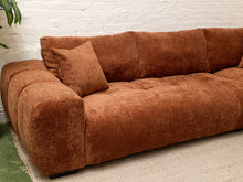 Load image into Gallery viewer, A modern sectional sofa upholstered in a rust-brown corduroy fabric, featuring a matching throw pillow, set against a white brick wall and a light-colored rug.
