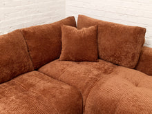 Load image into Gallery viewer, A close-up of a corner sectional sofa upholstered in rust-brown corduroy fabric, with matching throw pillows, set against a white brick wall.
