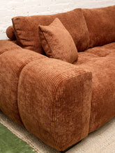 Load image into Gallery viewer, A close-up of a modern sectional sofa upholstered in a rust-brown corduroy fabric, featuring a matching throw pillow, set against a white brick wall background.
