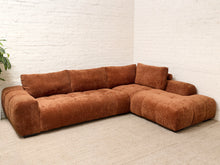 Load image into Gallery viewer, A modern L-shaped sectional sofa upholstered in a textured rust-brown bouclé fabric, set against a white brick wall and light beige carpet.
