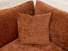 Load image into Gallery viewer, A rust-brown corduroy throw pillow centered on a matching corduroy sofa, with two larger matching back cushions behind it against a white painted brick wall.
