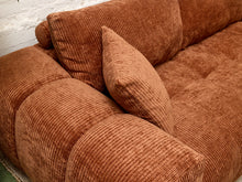 Load image into Gallery viewer, A close-up of a chunky, barrel-style sofa upholstered in a rust-brown corduroy fabric, featuring matching throw pillows against a white brick wall.
