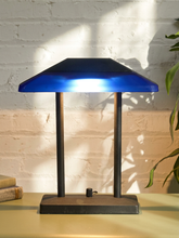 Load image into Gallery viewer, A modern table lamp with a vibrant blue glass shade and a dark metal base, shown lit on a table against a white brick wall with warm sunlight and leaf shadows.
