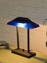 Load image into Gallery viewer, A modern table lamp with a glowing blue translucent shade and a dark metal base, shown on a tabletop against a white brick wall.
