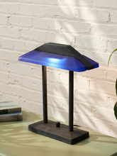 Load image into Gallery viewer, A modern table lamp with a geometric, house-shaped shade in blue and black, supported by two slim black legs on a square base, set on a table against a white brick wall.
