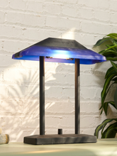 Load image into Gallery viewer, A modern table lamp with a dark charcoal base and two vertical support posts, featuring a translucent blue glass shade that glows with warm light from within, set against a white brick wall background.
