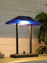 Load image into Gallery viewer, A modern table lamp with a dark metal frame and a translucent blue shade glowing with warm light, set against a white brick wall with a green plant in the background.
