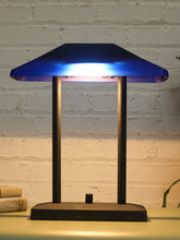 Load image into Gallery viewer, A modern table lamp with a dark charcoal base and two vertical support posts, featuring a translucent blue glass shade that glows with a cool blue light when turned on, set against a white brick wall background.
