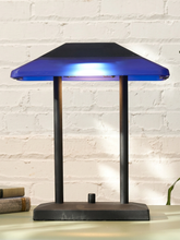Load image into Gallery viewer, A modern table lamp with a dark charcoal base and two vertical support posts, featuring a translucent blue glass shade that glows with cool white light. The lamp is set on a light-colored surface against a white brick wall background.
