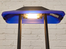Load image into Gallery viewer, A close-up of a portable LED light bar with a bright glowing bulb, mounted on a blue and black device with two black support legs, set against a white brick wall background.
