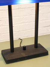 Load image into Gallery viewer, A vintage-style table lamp with a rectangular black base, two vertical black legs, and a bright blue conical shade, shown against a white brick wall background.
