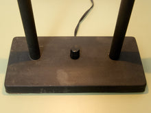 Load image into Gallery viewer, Close-up of the base of a black metal lamp stand with two vertical legs and a central round knob, with a power cord visible behind it on a light beige surface.
