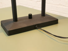 Load image into Gallery viewer, Close-up of a black metal lamp base with two vertical posts and a power cord plugged into a wall socket, resting on a light green surface.
