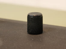 Load image into Gallery viewer, A small black ribbed cylindrical cap or knob standing upright on a dark surface with a soft beige background.
