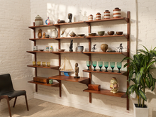 Load image into Gallery viewer, A mid-century modern wall-mounted wooden open shelving unit with multiple tiers, styled with various home decor items including vases, bowls, glasses, and a small model ship, set against a white brick wall.

