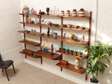 Load image into Gallery viewer, A set of mid-century modern floating wooden shelves in a warm medium-brown finish, styled with various home decor items, glassware, and a model sailboat, mounted on a white brick wall next to a green potted plant.
