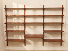 Load image into Gallery viewer, A mid-century modern wall-mounted shelving unit made of warm brown wood, featuring five open shelves and a lower console shelf, mounted on a white brick wall.
