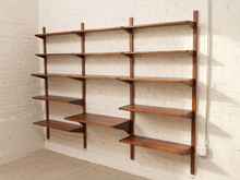 Load image into Gallery viewer, A mid-century modern wall-mounted wooden shelving unit with multiple tiers in a warm medium-brown finish, mounted on a white brick wall.
