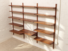 Load image into Gallery viewer, Mid-century modern wall-mounted wooden bookshelves in a warm medium-brown finish, mounted on a white brick wall above a beige carpeted floor.

