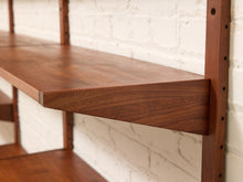 Load image into Gallery viewer, Close-up of a dark brown wooden floating shelf mounted on a white brick wall with matching vertical support brackets.
