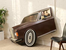 Load image into Gallery viewer, A vintage-style car-shaped room divider or play couch made of brown wood, leaning against a white brick wall in a room with a potted plant and a black chair.
