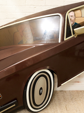 Load image into Gallery viewer, A close-up of a brown car interior and exterior, showing a person sitting in the driver's seat with a bright reflection on the window. The car features cream-colored trim and whitewall-style wheels.
