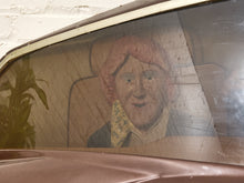 Load image into Gallery viewer, A close-up of a vintage brown car window showing a painted wooden figure or doll with pink hair and a patterned yellow neck scarf, set against a light wall.
