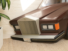 Load image into Gallery viewer, A vintage-style brown car front end displayed indoors on a beige carpet against a white brick wall, with a plant leaf visible at the left edge.
