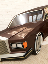 Load image into Gallery viewer, A close-up of a brown classic car model or toy car with chrome grille, white trim, and orange side marker lights, positioned against a white brick wall and beige carpet.
