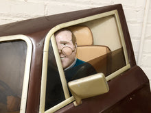Load image into Gallery viewer, A novelty car window prop with a vintage brown body, cream trim, and a realistic mannequin head sitting inside, set against a white brick wall.

