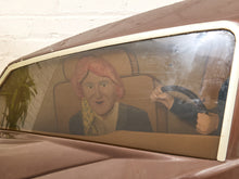 Load image into Gallery viewer, A vintage brown car window with a painted cardboard cutout of a person sitting in the back, photographed against a white brick wall.
