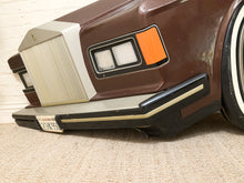 Load image into Gallery viewer, Close-up of the front corner of a vintage-style car with a brown body, cream grille and trim, black bumper, and an orange side marker light, photographed indoors against a light wall and carpet.
