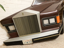 Load image into Gallery viewer, A close-up of a vintage-style car front end, featuring a large vertical slatted chrome grille, dual rectangular headlights, a silver bumper, and a California license plate, parked against a white brick wall.

