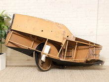 Load image into Gallery viewer, A partially disassembled vintage car body panel with exposed wooden frame, black metal chassis, and a single wheel, photographed indoors against a white brick wall.
