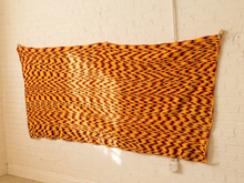Load image into Gallery viewer, A large rectangular textile with an orange, yellow, and black wavy abstract pattern hanging on a white brick wall.
