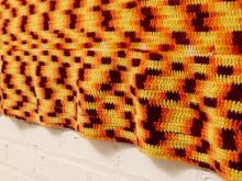 Load image into Gallery viewer, A close-up of a textured crochet blanket or throw featuring a geometric pattern in orange, yellow, and dark brown, draped over a white brick wall.
