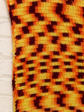 Load image into Gallery viewer, A close-up of a textured crochet or knit fabric with a geometric pattern in orange, yellow, and dark brown, shown against a white brick wall background.
