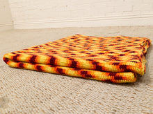 Load image into Gallery viewer, A folded chunky knit blanket with a lava pattern in yellow, orange, and dark red, resting on a textured beige rug against a white brick wall.
