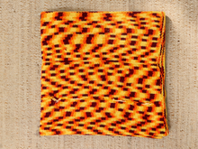 Load image into Gallery viewer, A folded square woven textile with a bold zigzag optical illusion pattern in orange, yellow, and black, displayed flat on a textured beige fabric background.
